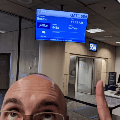 A man in an airport, looking and pointing up to a monitor that shows the gate number, time and location for a flight.