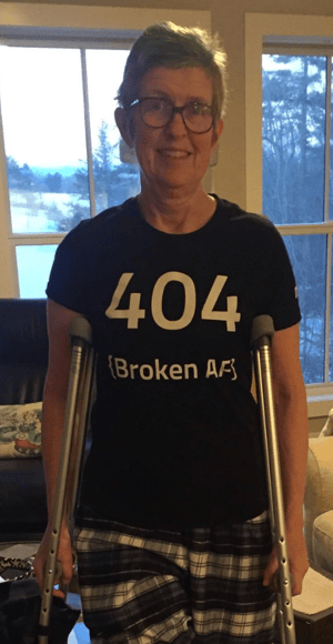 A woman standing with crutches in a living room. She is wearing a black t shirt that says 404 {Broken AF}